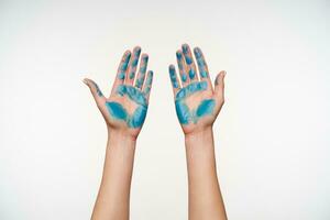 Front view of lady's hands painted with blue colour being raised while demonstrating palms, posing over white background. Human hands concept photo