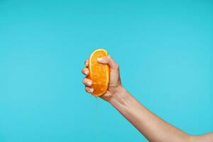 Indoor photo of young attractive hand holding orange and clenching a fists while queezing juice, preparing breakfast while posing over blue background