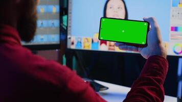 Photo editor using green screen mobile phone to watch tutorial, learning to color correct photographs in creative studio. Photographer watching guiding video on chroma key smartphone