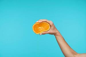 Beautiful fair-skinned pretty hand keeping her hand raised while holding fresh orange, squeezing it with fingers while isolated against blue background photo