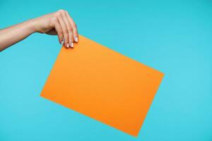 Young secretary with white manicure keeping her hand raised while holding orange envelope with important documents while standing over blue background photo