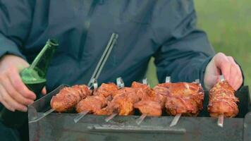 man är matlagning fläsk kött utomhus, innehav öl i grön flaska och vändning utegrill på spett. varm grillad kebab på de kol. picknick mat förbereder. mitten närbild skott video