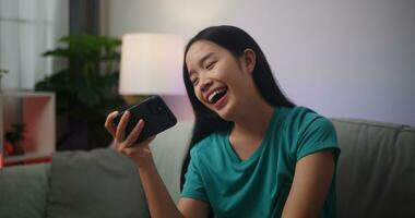 retrato de joven asiático mujer disfruta jugando en línea deporte juegos en teléfono inteligente sentado en sofá en el vivo habitación a hogar, jugador estilo de vida concepto. foto