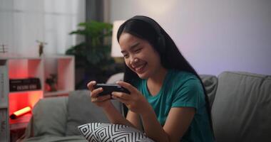 retrato de joven asiático mujer vistiendo lentes y auriculares disfruta jugando en línea deporte juegos en teléfono inteligente sentado en sofá en el vivo habitación a hogar, jugador estilo de vida concepto. foto