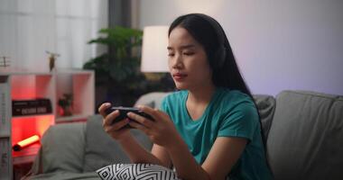 retrato cerca arriba de joven asiático mujer vistiendo lentes y auriculares disfruta jugando en línea deporte juegos en teléfono inteligente sentado en sofá en el vivo habitación a hogar, jugador estilo de vida concepto. foto