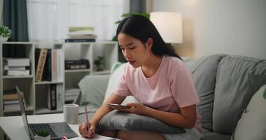 Portrait of Young Asian woman checks her finances using a calculator on smartphone and makes notes in a notebook while sitting on the sofa at home,Home finance photo