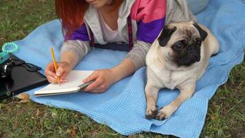 Girl laying on a lawn and writing, her pug laying beside video