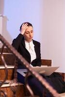 Overworked tired businesswoman with headache working on difficult project deadline. Serious entrepreneur working on job project sitting on staircase of business building at night hours for job. photo