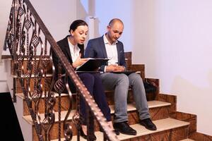 Worried tired businessman doing overtime at night on job diffuclt deadline with coworker sitting on stairs. Colleague entrepreneur working late together in evening at corporate job sitting on staircase, explaining difficult finance project. photo