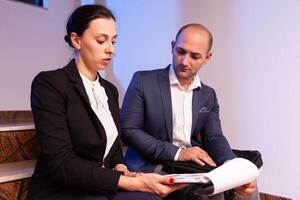 Overworked businessman reading raport after hours about deadline financial crisis late at night with coworker. Colleague entrepreneur working late together in evening at corporate job sitting on staircase, explaining difficult finance project. photo