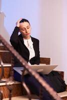 Overworked exhausted businesswoman falling asleep working on project deadline. Serious entrepreneur working on job project sitting on staircase of business building at night hours for job. photo