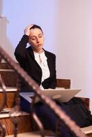 Stressed overworked businesswoman doing overtime for project deadline at late hours. Serious entrepreneur working on job project sitting on staircase of business building at night hours for job. photo