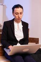 Stressed overworked businesswoman using notebook for business deadline late in the evening. Serious entrepreneur working on job project sitting on staircase of business building at night hours for job. photo