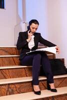 Stressed tired businesswoman readling project deadline during conversation on smartphone. Serious entrepreneur working on job project sitting on staircase of business building at night hours for job. photo
