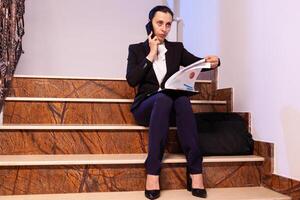 Overworked tired businesswoman reading project deadline during phone call with coworker. Serious entrepreneur working on job project sitting on staircase of business building at night hours for job. photo