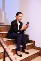 Stressed exhausted overworked businesswoman working on job deadling using smartphone overtime. Serious entrepreneur working on job project sitting on staircase of business building at night hours for job. photo