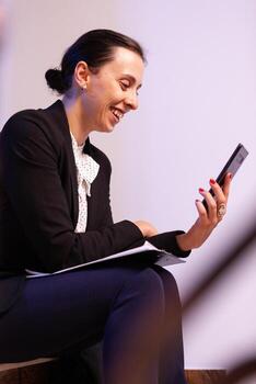 Overworked businesswoman smiling during video call about project deadline. Entrepreneur talking with client using smartphone for video call sitting on stairs. photo