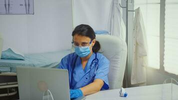 Nurse typing on laptop wearing medical gloves during coronavirus. Physician doctor specialist in medicine providing health care services consultation treatment examination in hospital cabinet photo