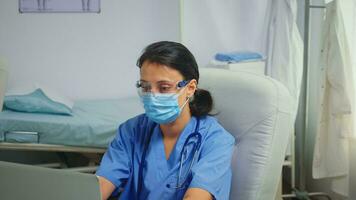 Ward assistant having protection mask and glasses writing a report on laptop. Physician specialist in medicine providing health care services consultation treatment in hospital cabinet during covid19 photo