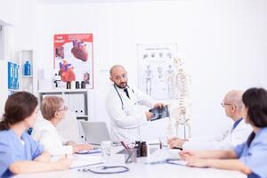 Surgeon holding radiography and making presentation in front of colleagues in hospital meeting room. Clinic expert therapist talking with coworkers about disease, medicine professional photo