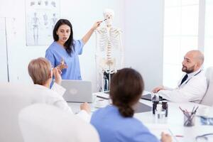 Female radiologist doctor holding presentation using skeleton in front of medical staff. Clinic expert therapist talking with colleagues about disease, medicine professional photo