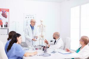 Mature doctor wearing glasses while holding presentation about human anatomy using skeleton. Clinic expert therapist talking with colleagues about disease, medicine professional photo