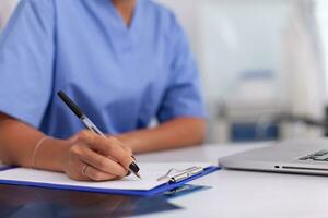 Close up of nurse writing patient diagnosis on clipboard in hospital office . Health care physician using computer in modern clinic looking at monitor, medicine, profession, scrubs. photo