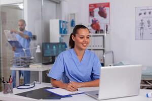 Smiling nurse using laptop computer and writing notes on clipboard in hospital office with doctor in the background. Health care physician using computer in modern clinic looking at monitor, photo