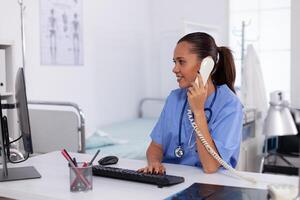 médico enfermero hablando con paciente en el teléfono acerca de diagnóstico. salud cuidado médico sentado a escritorio utilizando computadora en moderno clínica mirando a monitor. foto