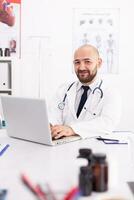 Portrait of doctor smiling holding hands on laptop keyboard looking at camera in hospital office wearing lab coat. Medical practitioner using notebook in clinic workplace, expertise, medicine. photo