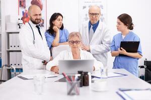 Mature doctor specialist briefing her medical team in conference room using laptop. Clinic expert therapist talking with colleagues about disease, medicine professional photo