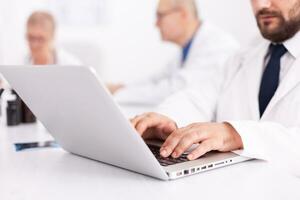 Young medical specialist typing on laptop during meeting with coworkers in hospital office. Clinic expert therapist talking with colleagues about disease, medicine professional photo