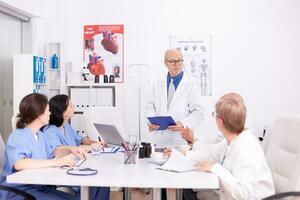 Expert elderly aged male doctor having a discussion with medical staff in hospital conference room. Clinic expert therapist talking with colleagues about disease, medicine professional photo