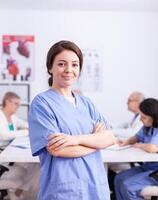 Medical therapist looking at camera with healthcare staff in the background wearing uniform. Friendly medical practitioner in clinic meeting room, robe, specialist. photo