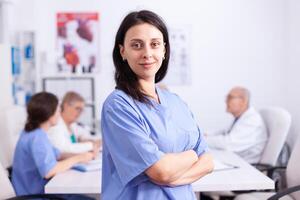 Smiling nurse in wearing blue uniform in hospital conference room looking at camera with medical staff in the background. Friendly medical practitioner in clinic meeting room, robe, specialist. photo