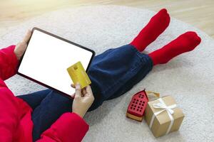 A woman shopping online sitting on carpet at home with a tablet with a white screen isolated using a gold credit card. Use your credit card to tap the screen to verify your identity. Clipping Path. photo