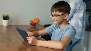 das Kind Verwendet das Tablette während Sitzung im das Küche während Mama ist vorbereiten Frühstück. video