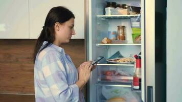 ein Frau ist Planung ein Essen aufführen in der Nähe von das Kühlschrank mit ein Handy, Mobiltelefon App. video