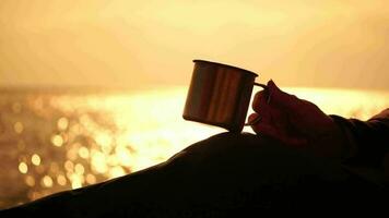 Travel stainless steel mug in the hands of a woman. Small waves on golden warm water surface with bokeh lights from sun. A concept of the world of beauty, nature and outdoor travel. Close up video