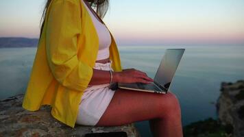 digital nômade, mulher dentro a chapéu, uma o negócio mulher com uma computador portátil senta em a pedras de a mar durante pôr do sol, faz uma o negócio transação conectados a partir de uma distância. freelance, controlo remoto trabalhos em período de férias. video