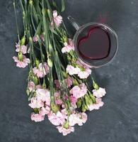 pink carnations on a dark background with a cup of red tea in a heart-shaped cup photo