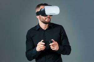 emocional joven hombre utilizando un vr auriculares y experimentando virtual realidad en gris antecedentes foto