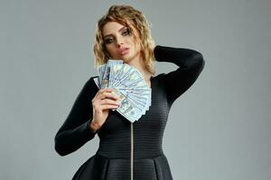 Blonde maiden with curly hair, in black dress is holding some cash, posing on gray background. Gambling entertainment, poker, casino. Close-up photo