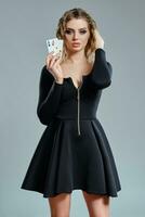 Blonde lady in black short dress showing two playing cards, posing against gray background. Gambling entertainment, poker, casino. Close-up. photo