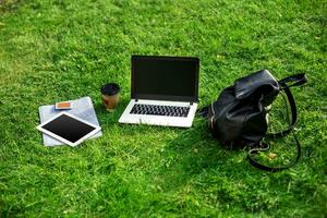 Laptop computer on green grass with coffee cup, bag and tablet in outdoor park photo