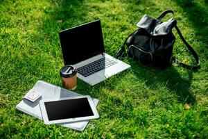 Laptop computer on green grass with coffee cup, bag and tablet in outdoor park photo