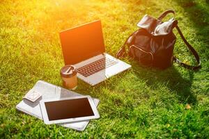 Laptop computer on green grass with coffee cup, bag and tablet in outdoor park photo