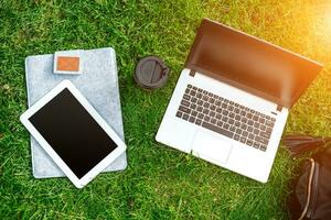 Laptop computer on green grass with coffee cup, bag and tablet in outdoor park photo