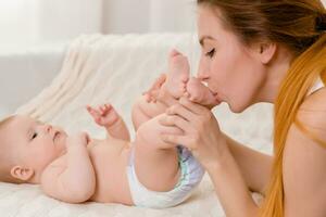 madre y niño en un blanco cama. mamá y bebé niña en pañal jugando en soleado dormitorio. foto