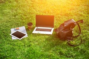 Laptop computer on green grass with coffee cup, bag and tablet in outdoor park photo
